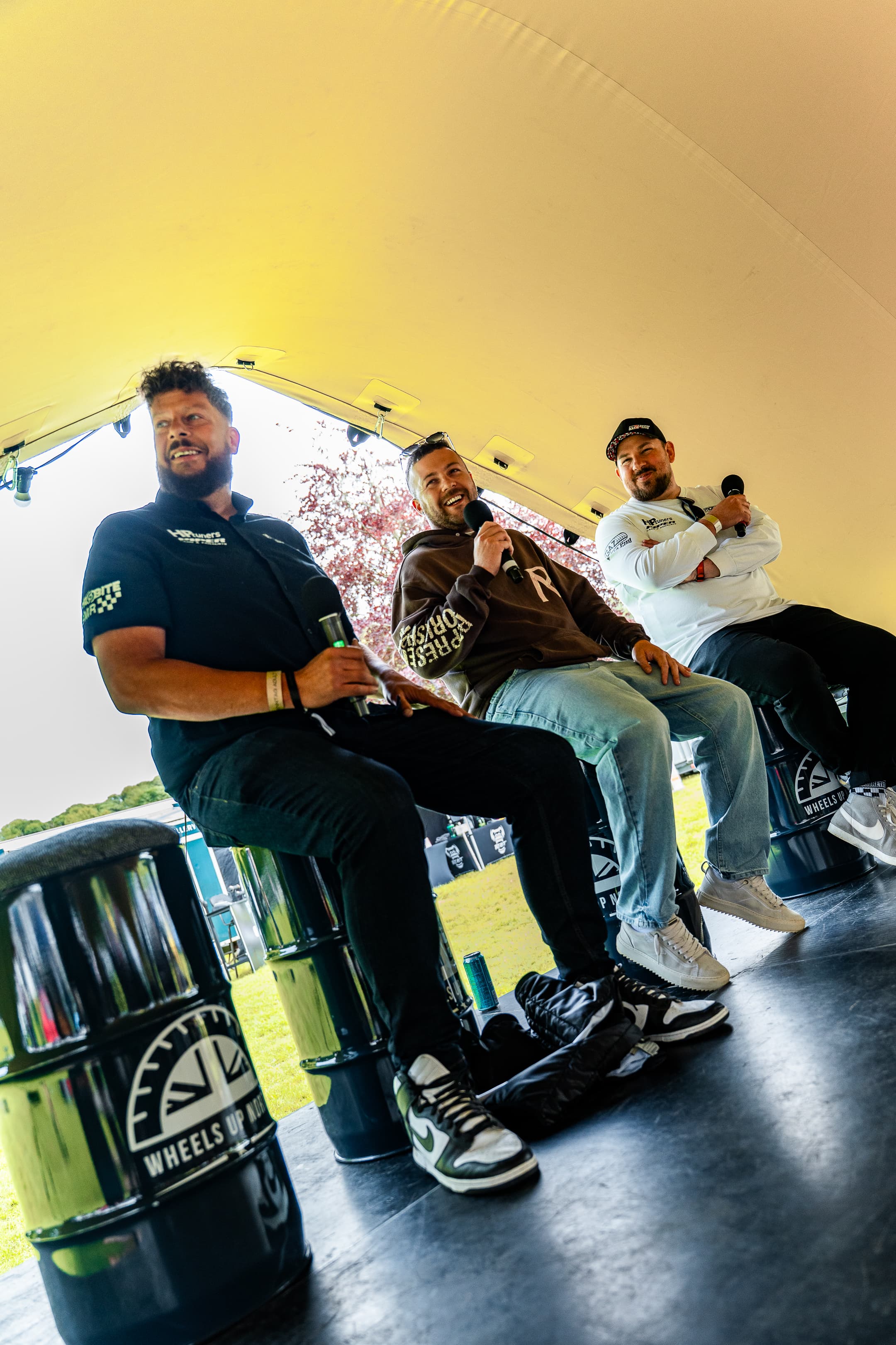 Three men chatting on a podcast panel, seated on branded oil-drum stools under a marquee