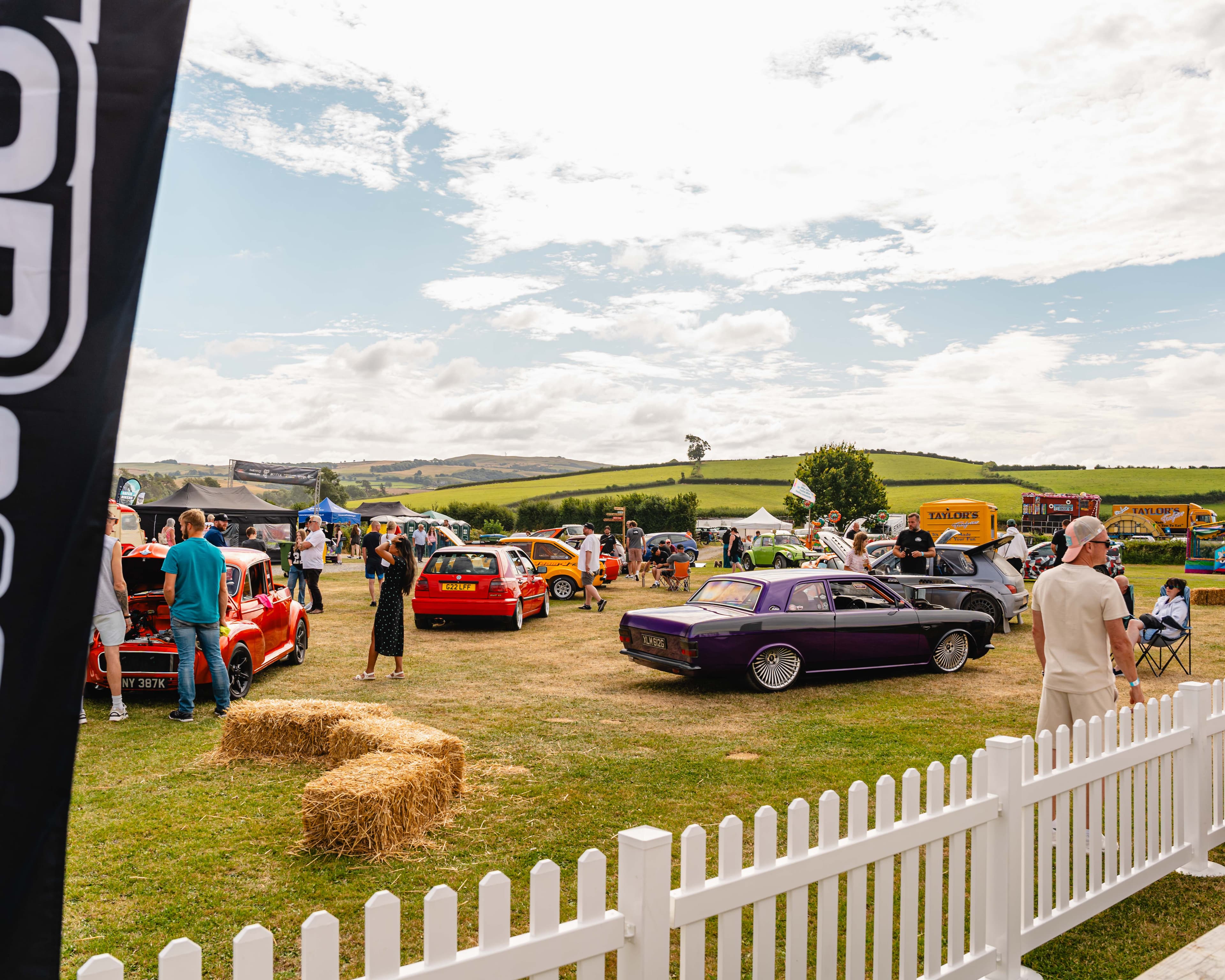 A wide view of a WUN car show field with modified cars, hay bales and rolling countryside in the background